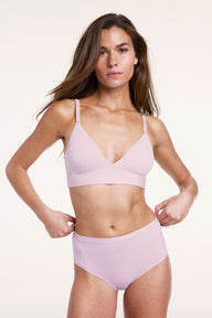 In a studio setting with a neutral backdrop, Gianna wears the Bliss Triangle Bralette and Bliss Highwaist Brief in Retro Lilac. She gently adjusts the waistband, highlighting the high-rise fit, full coverage, and ultra-soft microfiber fabric designed for everyday comfort. 