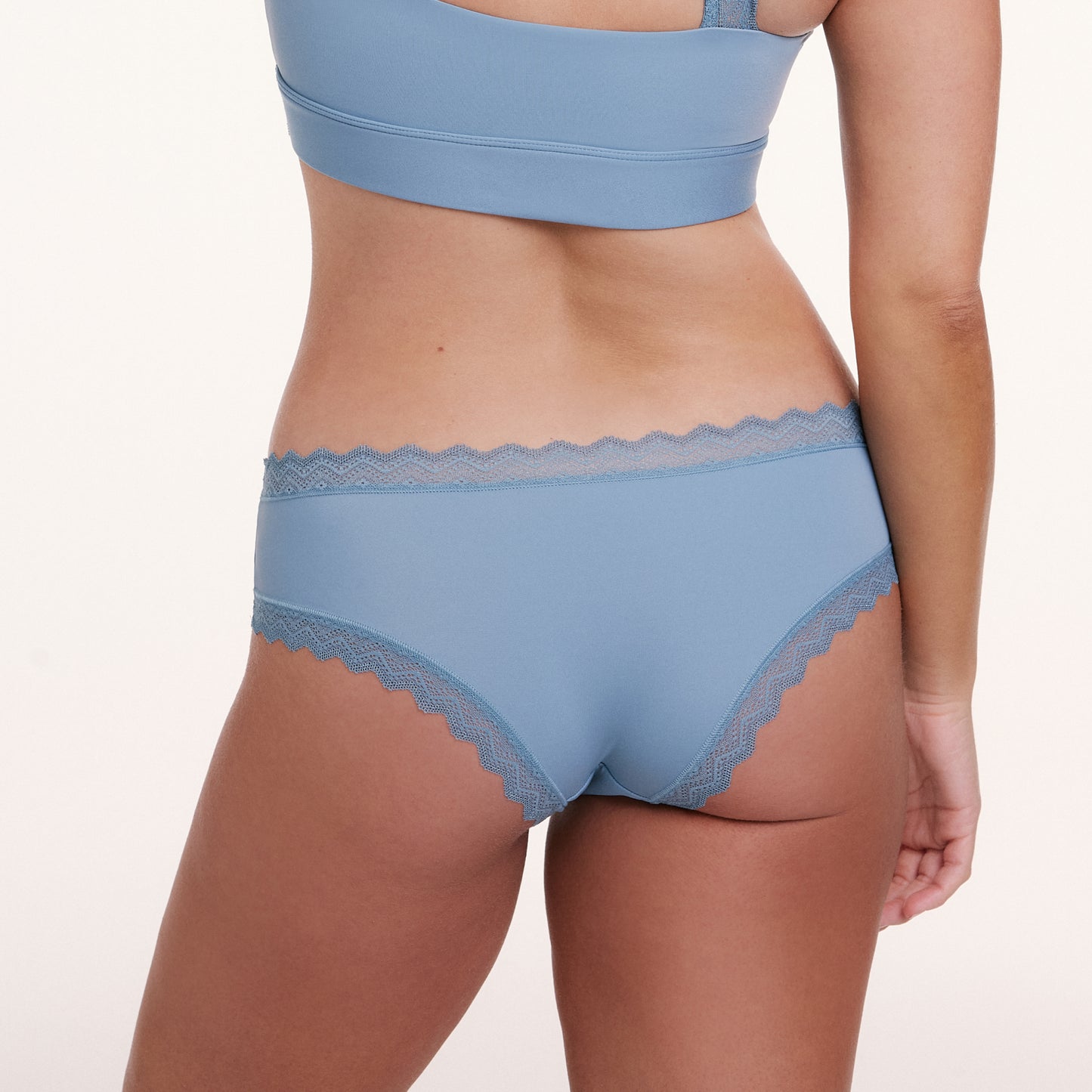 In a white studio setting, Sonnie wears the Bliss Lace Cheeky in Poolside, shown from the back with soft microfiber fabric and scalloped lace trim along the waistband and leg openings.