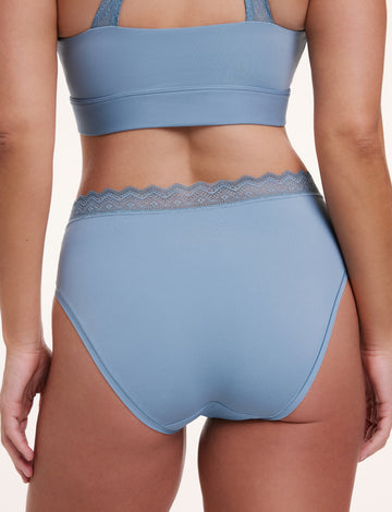 In a studio setting with a neutral backdrop, Sonnie wears the Bliss Lace High Waist Brief in Poolside blue, shown from the back. The brief features a high-rise fit with full back coverage and a scalloped lace waistband over ultra-soft microfiber fabric.