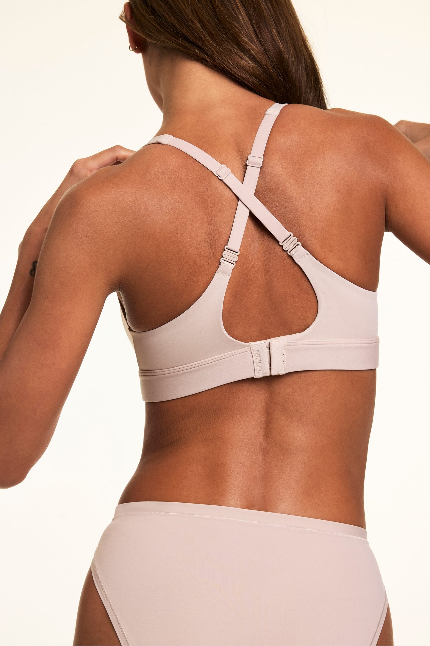 In a back studio close-up, Linni wears the Bliss Plunge Adjustable Bralette in Pebble with the straps crossed into a racerback style, showing the convertible straps and hook-and-eye back closure.