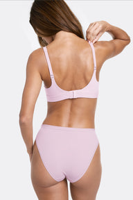 In a studio setting with a light gray backdrop, Linni wears the Bliss Plunge Adjustable Bralette in Retro Lilac, shown from the back while adjusting the strap. Thin adjustable straps, a hook-and-eye closure, and smooth microfiber fabric provide a customizable, comfortable fit.