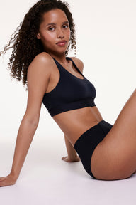 In a studio setting, Courtney wears the Bliss Scoop Bralette in Black with matching underwear, shown seated and highlighting the scoop neckline, wireless lift, and soft microfiber fabric for all-day comfort.