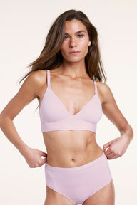 In a studio setting with a neutral backdrop, Gianna wears the Bliss Triangle Bralette and matching underwear in Retro Lilac. She gently adjusts the waistband, showing the deep V neckline, wireless band for lift, and smooth, ultra-soft microfiber fabric for everyday comfort. 