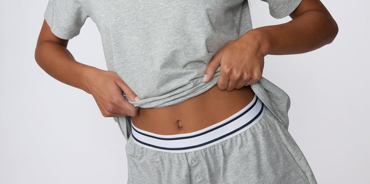 Against a clean white studio backdrop, a model lifts a grey tee to reveal Cloud Cotton sleep bottoms in Heather Grey, showing the soft elastic waistband with contrast stripe detailing and breathable cotton-modal fabric.