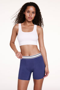 On a white studio backdrop, Courtney wears the Cloud Cotton Boxer Brief in Desert Shadow with a white bralette, gently holding the waistband. The boxer brief features a mid-rise waistband, longer inseam, and soft cotton modal fabric.