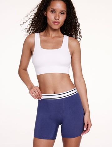 On a white studio backdrop, Courtney wears the Cloud Cotton Boxer Brief in Desert Shadow with a white bralette, gently holding the waistband. The boxer brief features a mid-rise waistband, longer inseam, and soft cotton modal fabric.