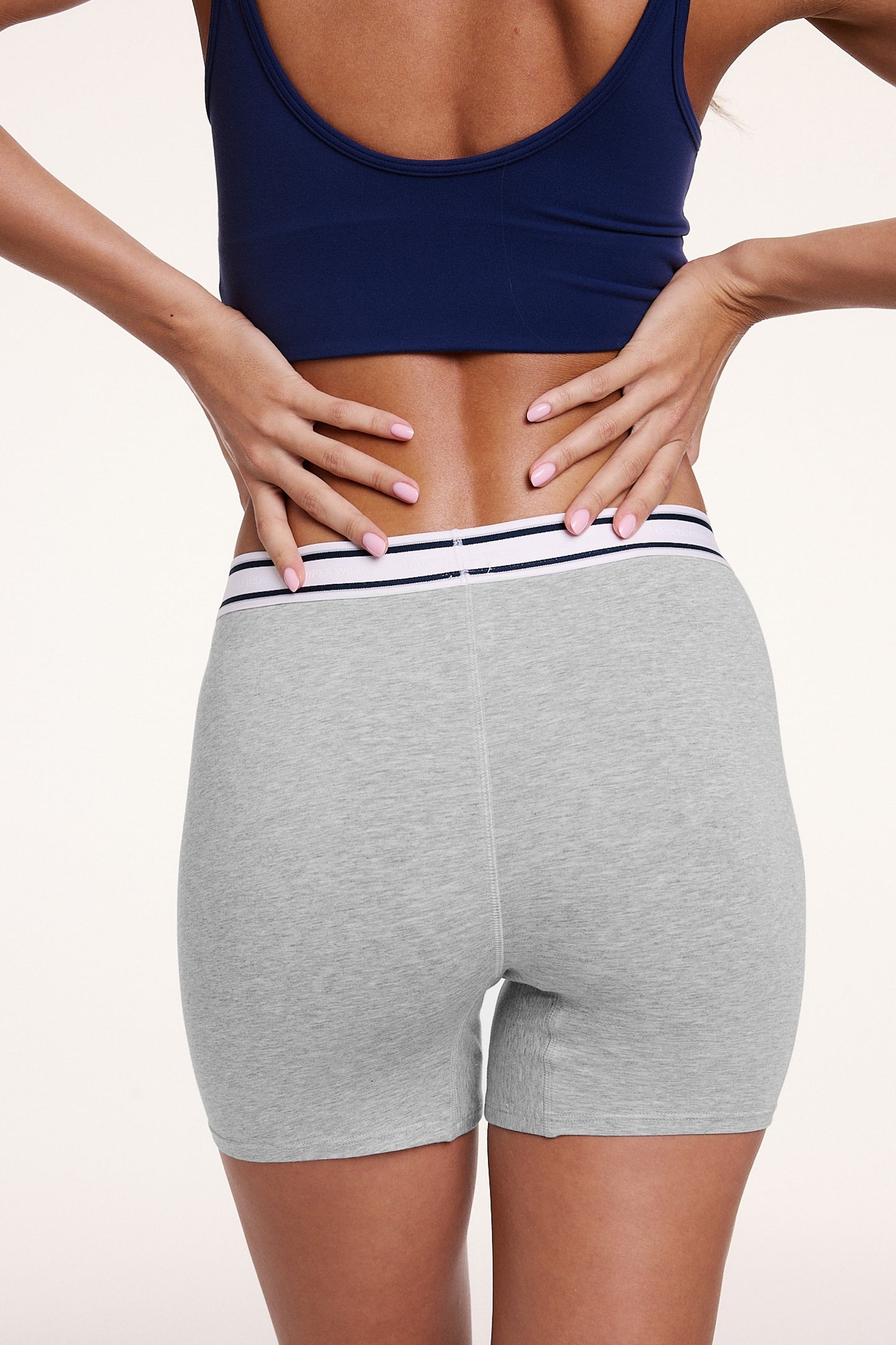 On a white studio backdrop, Linni wears the Cloud Cotton Boxer Brief in Heather Grey, shown from the back with hands at the waistband. The boxer brief offers full back coverage with a mid-rise fit and soft cotton modal fabric.
