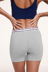 On a white studio backdrop, Linni wears the Cloud Cotton Boxer Brief in Heather Grey, shown from the back with hands at the waistband. The boxer brief offers full back coverage with a mid-rise fit and soft cotton modal fabric.