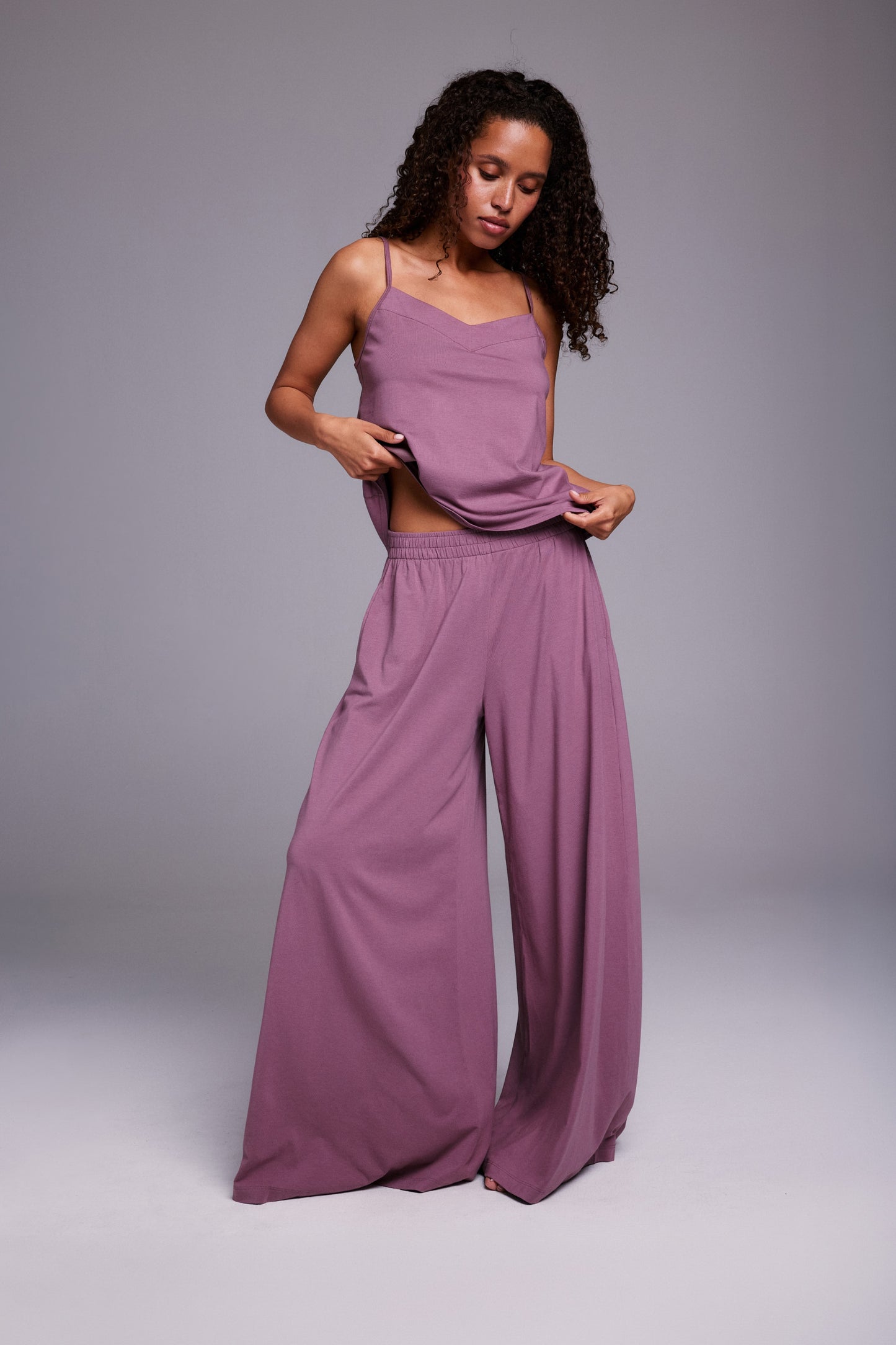 In a studio setting, Courtney wears a matching lounge set in Mirage, lifting the hem of a soft V-neck tank with thin straps to show the lightweight, breathable fabric and relaxed fit of the wide-leg pants.