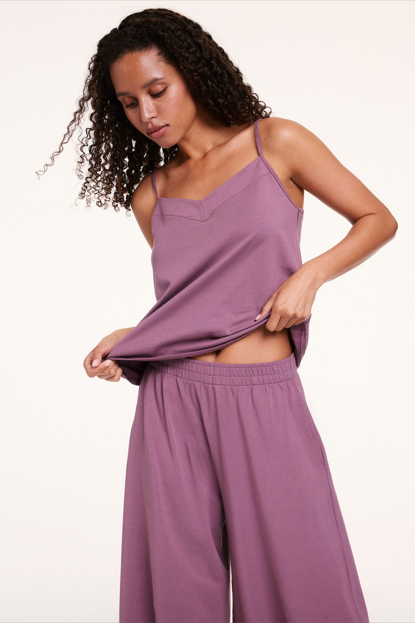 In a studio setting, Courtney wears a Cloud Cotton tank and pants in Mirage, lifting the hem to show the lightweight, breathable fabric and relaxed fit.