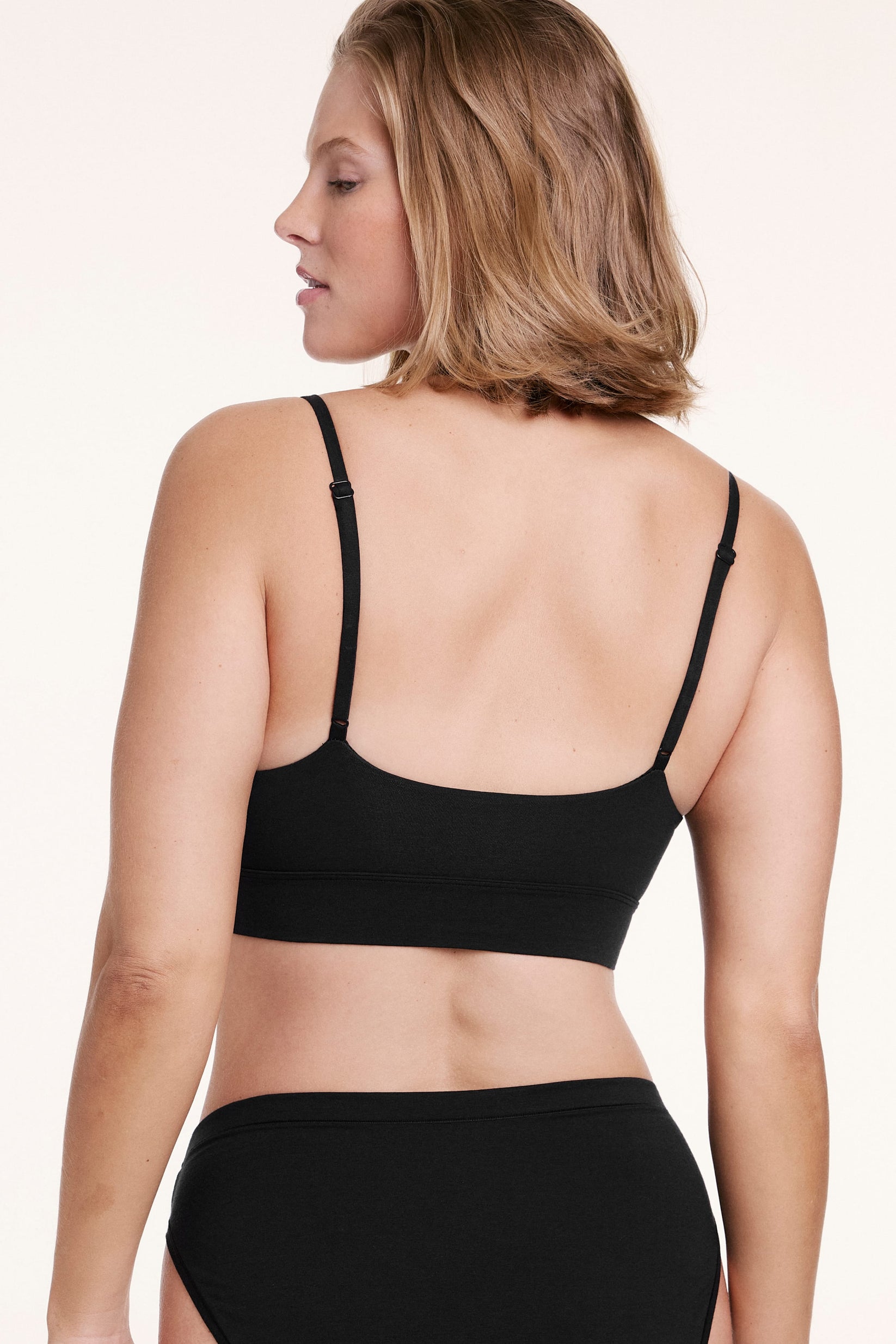 On a white studio backdrop, Sonnie wears the Cloud Cotton Triangle Bralette in Black, shown from the back with adjustable straps and a smooth, flexible band.