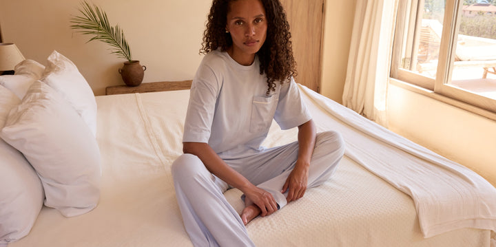 Model sitting cross-legged on a white bed in a sunlit, earth-toned bedroom, wearing the Harper Wilde Perfect Pima Pajama T-Shirt and matching wide-leg pant in Soft Blue.