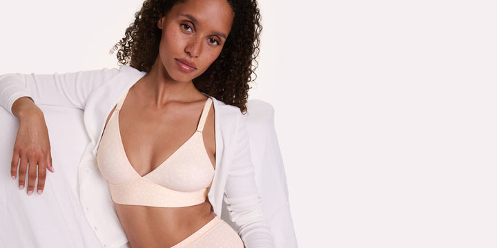 In a studio setting, Courtney wears the Bliss Triangle Bralette in Champagne Fizz with the Bliss Highwaist Brief in Champagne Fizz while seated in a white chair, highlighting the deep V neckline, front-adjusting straps, and soft microfiber fabric with a subtle dotted pattern.