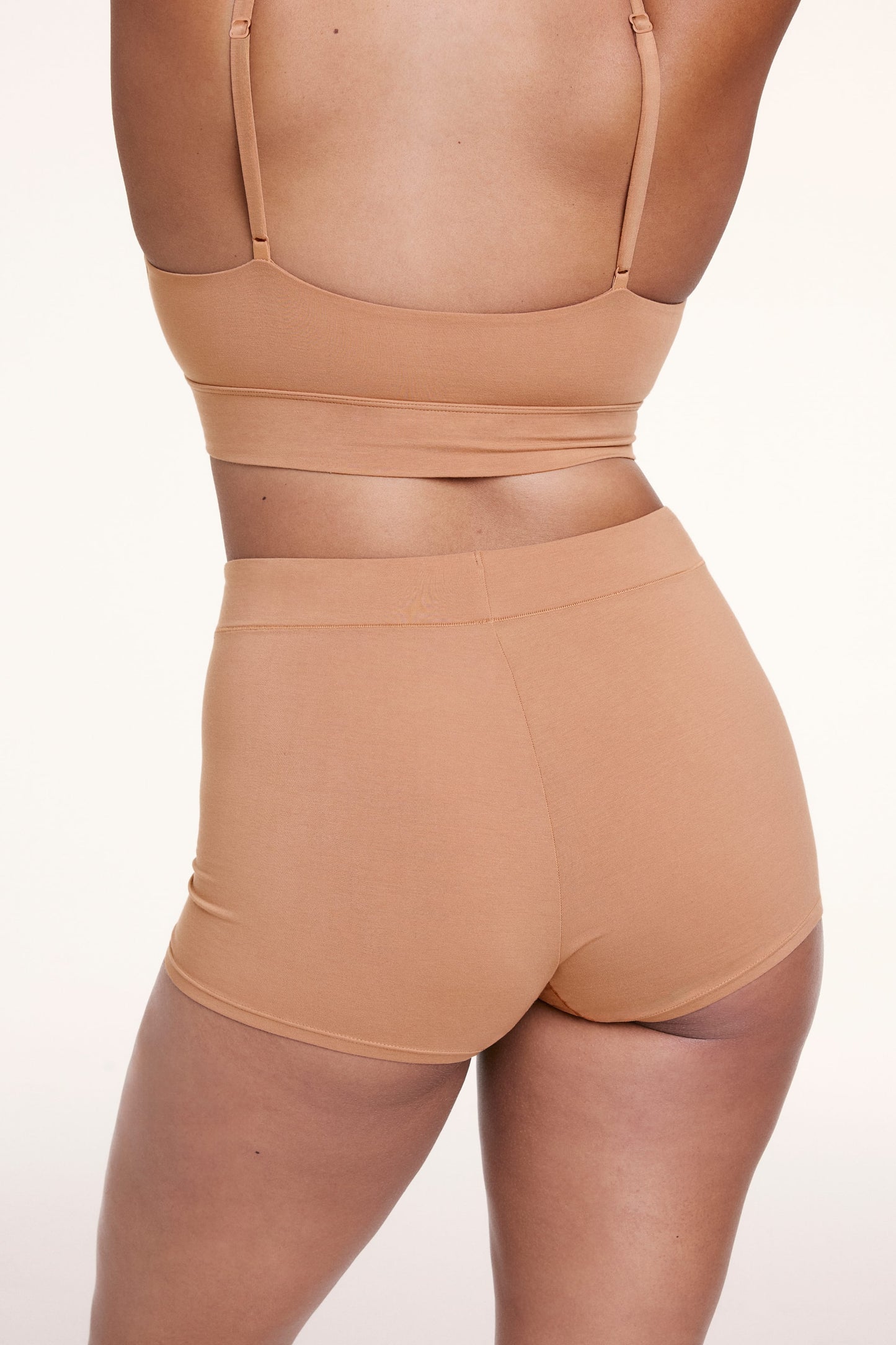 On a light studio backdrop, Alana wears the Cloud Cotton Boyshort in Dune, shown from the back. The boyshort features a high-rise waistband, full coverage, and a smooth, comfortable fit.