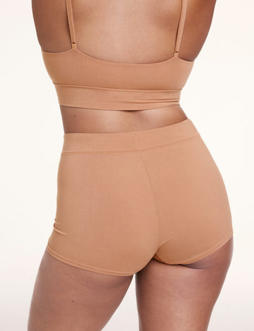 On a light studio backdrop, Alana wears the Cloud Cotton Boyshort in Dune, shown from the back. The boyshort features a high-rise waistband, full coverage, and a smooth, comfortable fit.