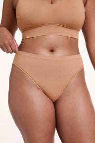 On a white studio backdrop, Alana wears the Cloud Cotton French Cut Brief in Dune, pulling slightly at the waistband to show the stretchy cotton modal fabric and high-rise fit. sizing: Alana wears size Large