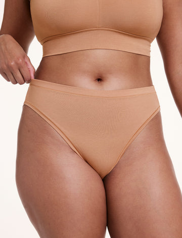 On a white studio backdrop, Alana wears the Cloud Cotton French Cut Brief in Dune, pulling slightly at the waistband to show the stretchy cotton modal fabric and high-rise fit. sizing: Alana wears size Large