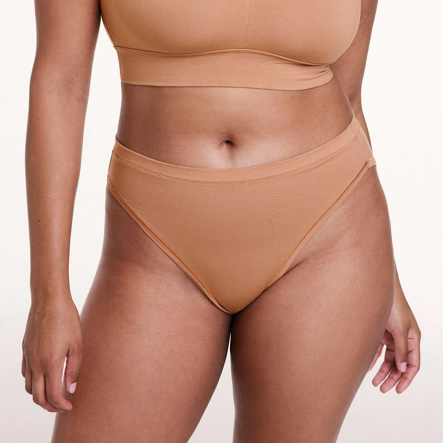 On a white studio backdrop, Alana wears the Cloud Cotton French Cut Brief in Dune, shown from the front with a high-rise waist and French cut leg opening.