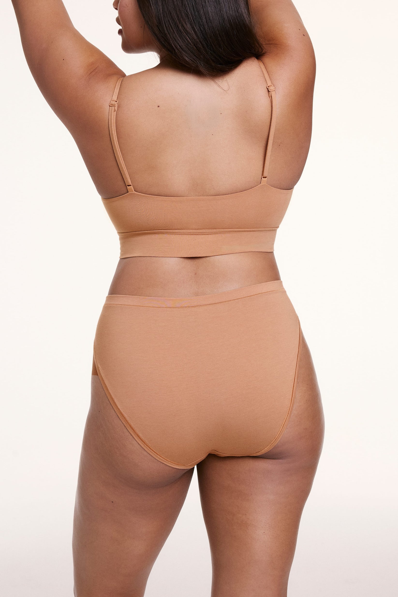 On a white studio backdrop, Alana wears the Cloud Cotton French Cut Brief in Dune with a matching bralette, shown from the back with mid- to full coverage and smooth, breathable fabric.