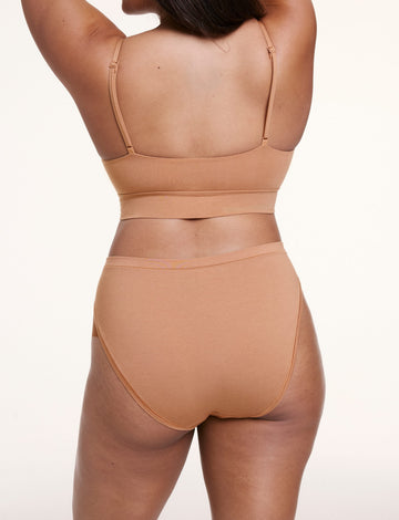 On a white studio backdrop, Alana wears the Cloud Cotton French Cut Brief in Dune with a matching bralette, shown from the back with mid- to full coverage and smooth, breathable fabric.