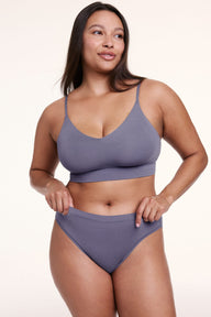 On a white studio backdrop, Alana wears the Cloud Cotton French Cut Brief in Slate with a matching bralette, holding the waistband to show the soft, stretchy cotton modal fabric.