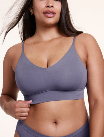 On a white studio backdrop, Alana wears the Cloud Cotton Triangle Bralette in Slate, shown from the front holding the underband to highlight the V neckline and soft, stretchy cotton modal fabric.  sizing: Alana wears size Large