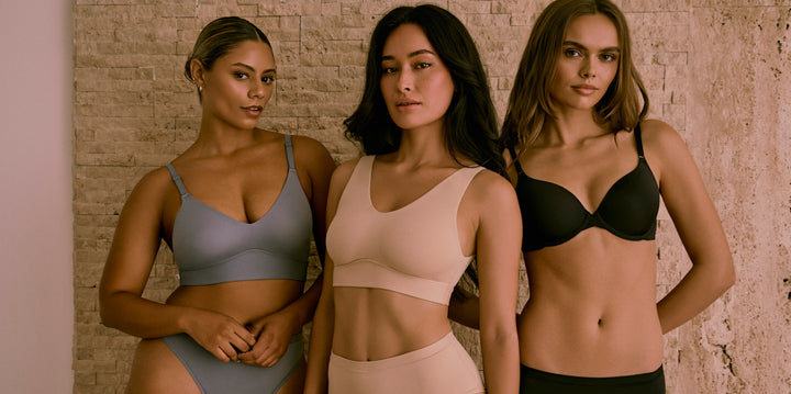 Three models pose in Harper Wilde Core Collection styles. From left to right: the first model wears the Bliss Triangle Bralette and Bliss Bikini in Steel; the second wears the Bliss Bralette and Bliss Highwaist Brief in Beige; the third wears the Base T-Shirt Bra and Bliss Hiphugger in Black. All stand confidently against a textured stone wall.