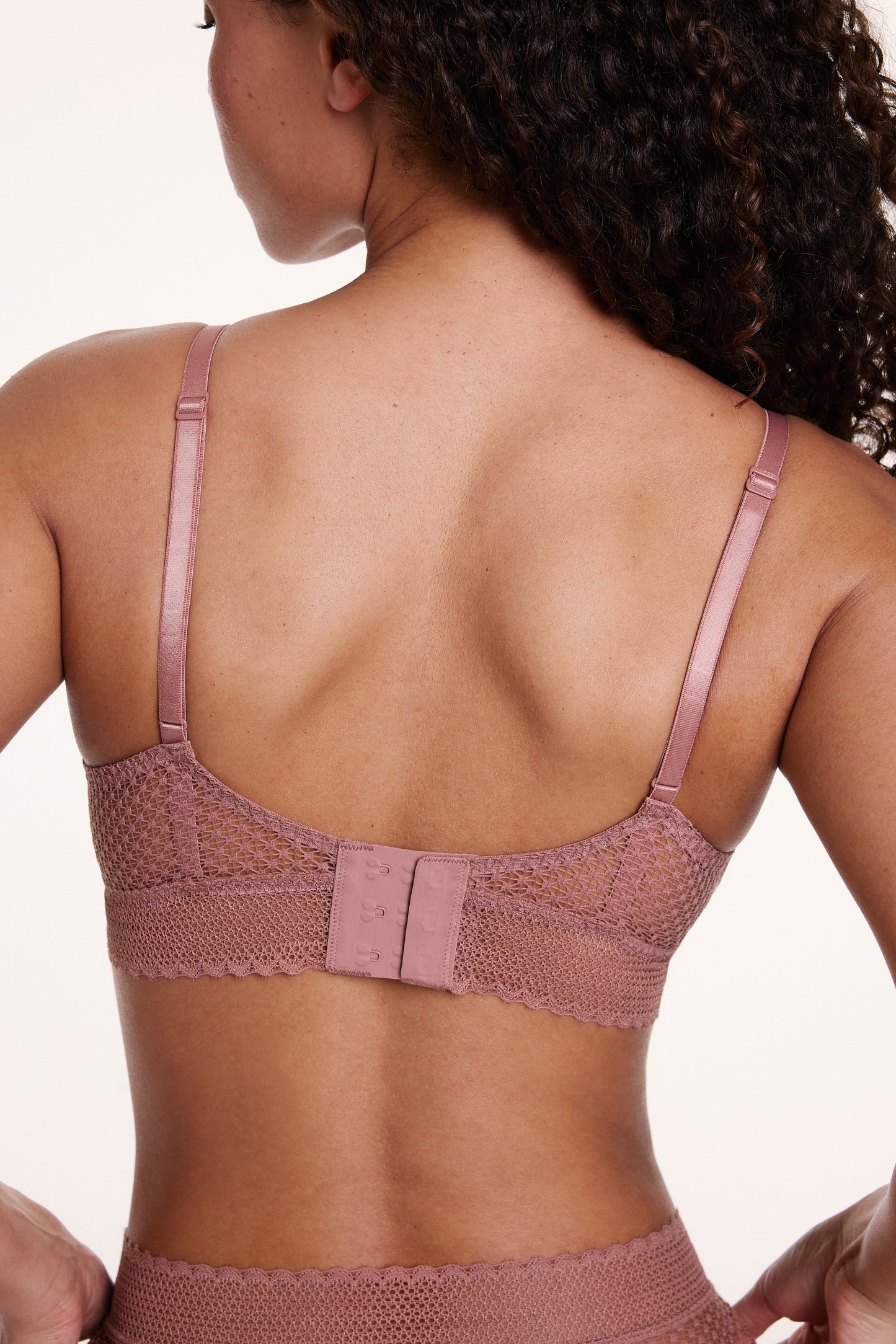 On a neutral studio backdrop, Courtney wears the Dot Lace Triangle Bralette in Plum from the back, showing the adjustable straps, sheer dot lace band, and hook-and-eye back closure.