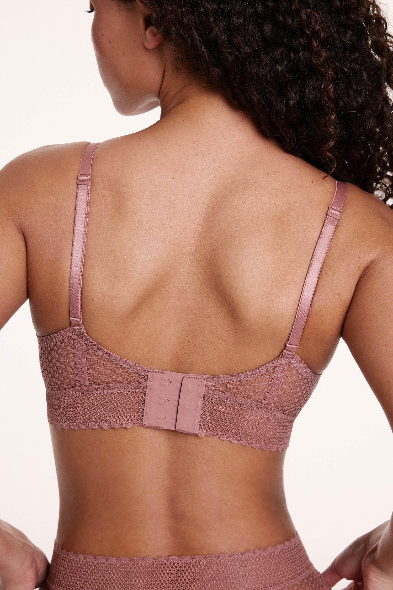 On a neutral studio backdrop, Courtney wears the Dot Lace Triangle Bralette in Plum from the back, showing the adjustable straps, sheer dot lace band, and hook-and-eye back closure.