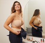 Summerlyn stands in front of a mirror in a well-lit room, wearing the Base Strapless Bra and No-Show Highwaist Brief in Beige. She is smiling and adjusting her navy dress, with makeup items visible on the counter, getting ready for an event.