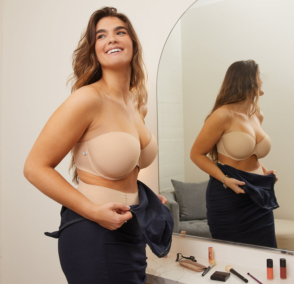 Summerlyn stands in front of a mirror in a well-lit room, wearing the Base Strapless Bra and No-Show Highwaist Brief in Beige. She is smiling and adjusting her navy dress, with makeup items visible on the counter, getting ready for an event.
