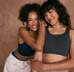 Two models are smiling and embracing against a warm, earthy backdrop. The model on the left is wearing the Lounge Rib Triangle Bralette in True Blue, while the model on the right is wearing the Lounge Rib Scoop Bralette in Charcoal.