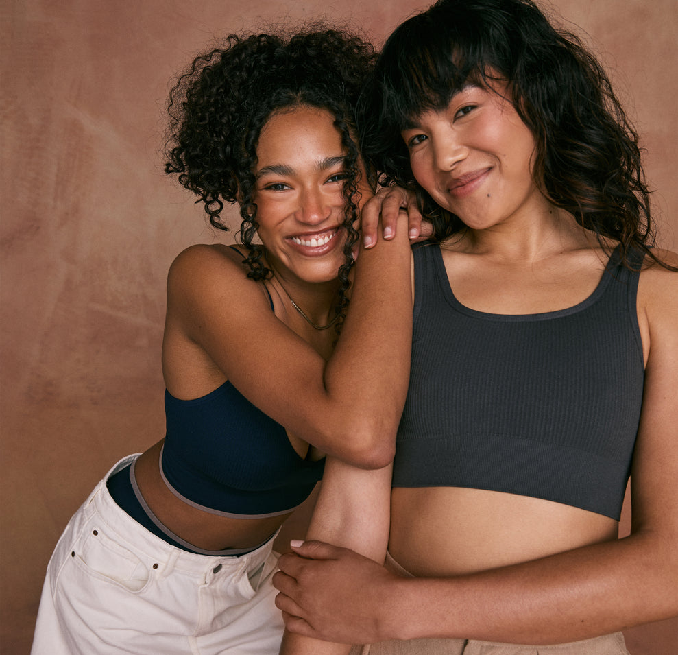 Two models are smiling and embracing against a warm, earthy backdrop. The model on the left is wearing the Lounge Rib Triangle Bralette in True Blue, while the model on the right is wearing the Lounge Rib Scoop Bralette in Charcoal.