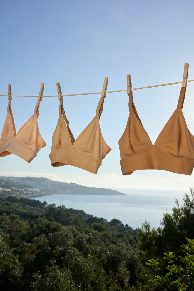 Three Bliss Triangle Bralettes in graduating nude shades hang from a sun-drenched clothesline against a bright blue sky and coastal Italian landscape.