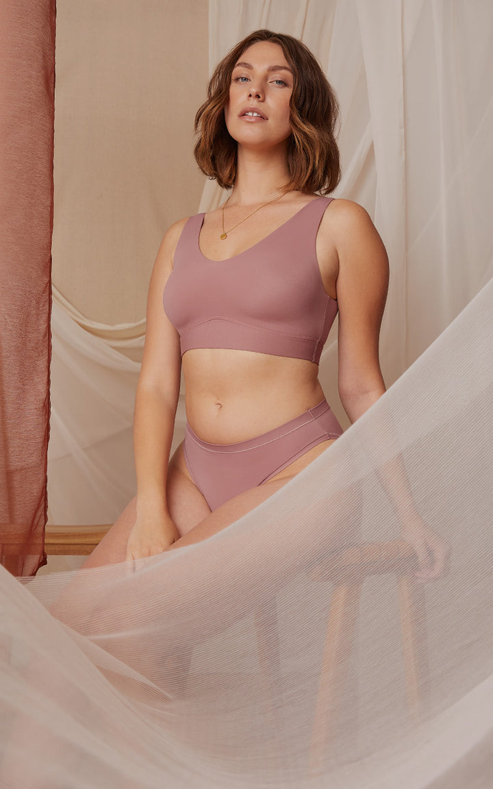 Three models pose in a softly lit setting with sheer fabric in the backdrop. The model on the left is wearing the Bliss Scoop Bralette and Bliss Highwaist Brief in Periwinkle. The model in the center is wearing the Bliss Triangle Bralette in Opal and Bliss Boyshort in Periwinkle. The model on the right is wearing the Bliss Bralette and Bliss Bikini in Plum.