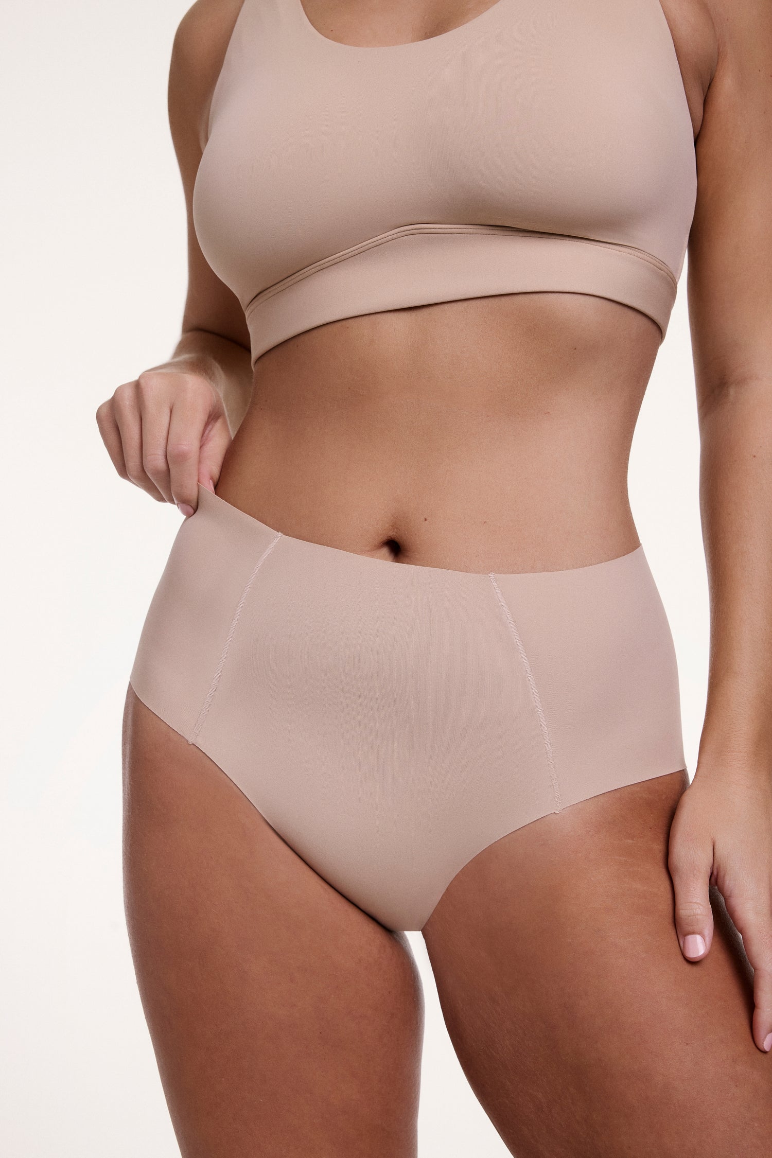On a white studio backdrop, Sonnie wears the No-Show Highwaist Brief in Beige, shown from the front with a smooth, high-rise waistband and raw-edge finish designed to sit flat against the skin. sizing: Sonnie wears size Large