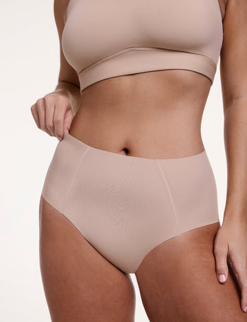 On a white studio backdrop, Sonnie wears the No-Show Highwaist Brief in Beige, shown from the front with a smooth, high-rise waistband and raw-edge finish designed to sit flat against the skin. sizing: Sonnie wears size Large