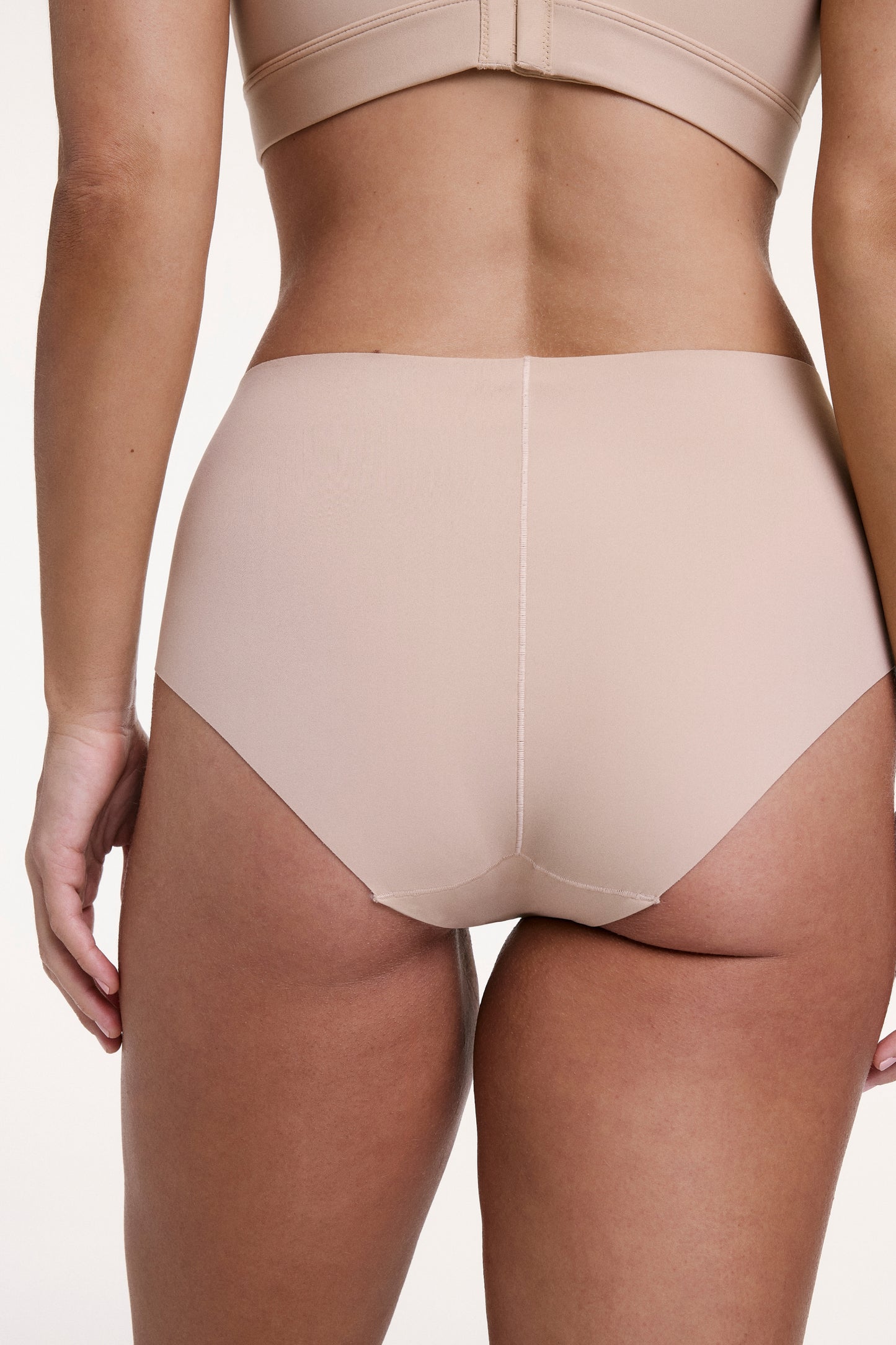 On a white studio backdrop, Sonnie wears the No-Show Highwaist Brief in Beige, shown from the back with full coverage and a smooth, raw-edge finish that lays flat under clothing.
