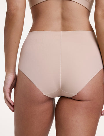 On a white studio backdrop, Sonnie wears the No-Show Highwaist Brief in Beige, shown from the back with full coverage and a smooth, raw-edge finish that lays flat under clothing.