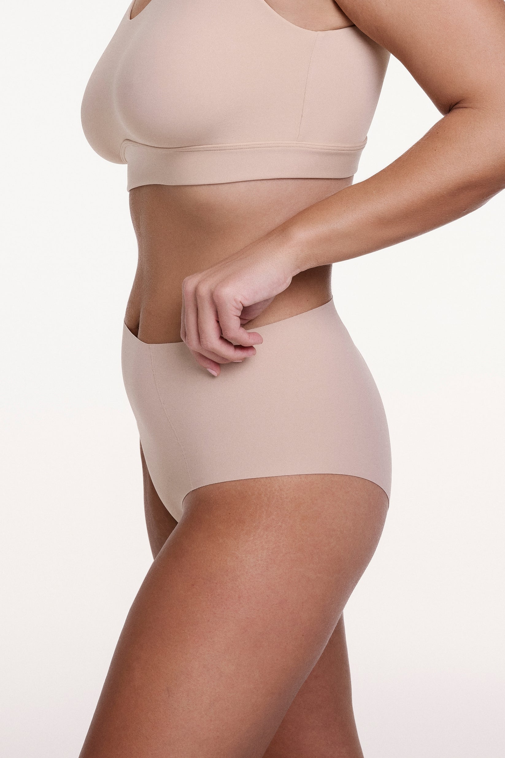 On a white studio backdrop, Sonnie wears the No-Show Highwaist Brief in Beige, shown from the side with a high-rise fit and smooth, elastic-free edges designed for all-day comfort.