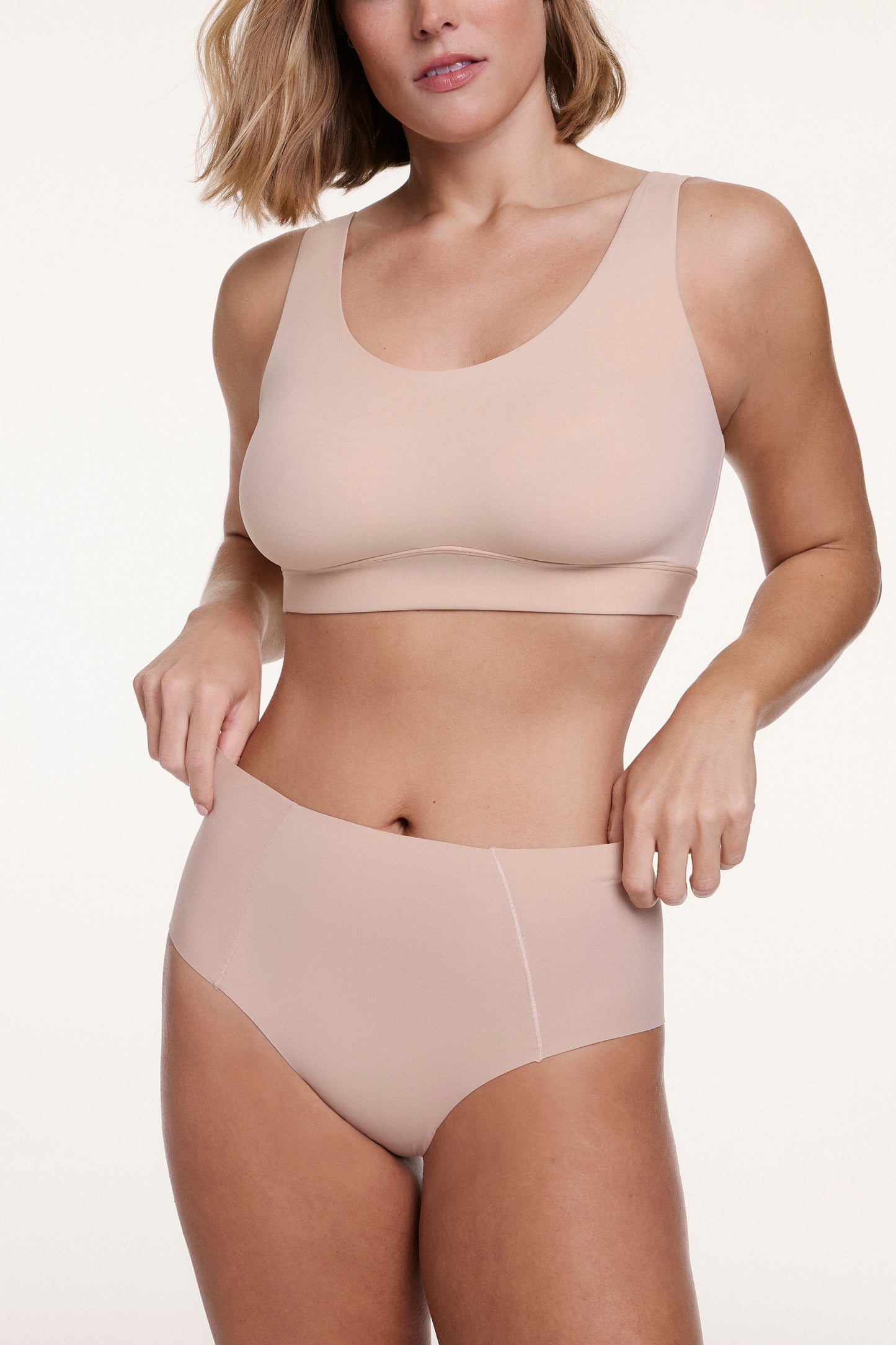 On a white studio backdrop, Sonnie wears the No-Show Highwaist Brief in Beige with a matching bra, gently pulling at the waistband to show the soft, stretchy fabric and high-rise fit.
