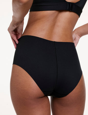 On a white studio backdrop, Courtney wears the No-Show High Waist Brief in Black, shown from the back with full coverage, a high-rise fit, and a smooth, seamless finish designed to lay flat under clothing. sizing: Courtney wears size Small