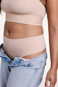 In a studio setting, Alana wears the No-Show Highwaist Thong in Beige with low-rise denim, showing the high-rise waistband and smooth, elastic-free edges designed to stay invisible under clothing. sizing: Alana wears size Large