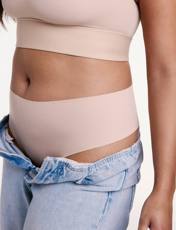 In a studio setting, Alana wears the No-Show Highwaist Thong in Beige with low-rise denim, showing the high-rise waistband and smooth, elastic-free edges designed to stay invisible under clothing. sizing: Alana wears size Large
