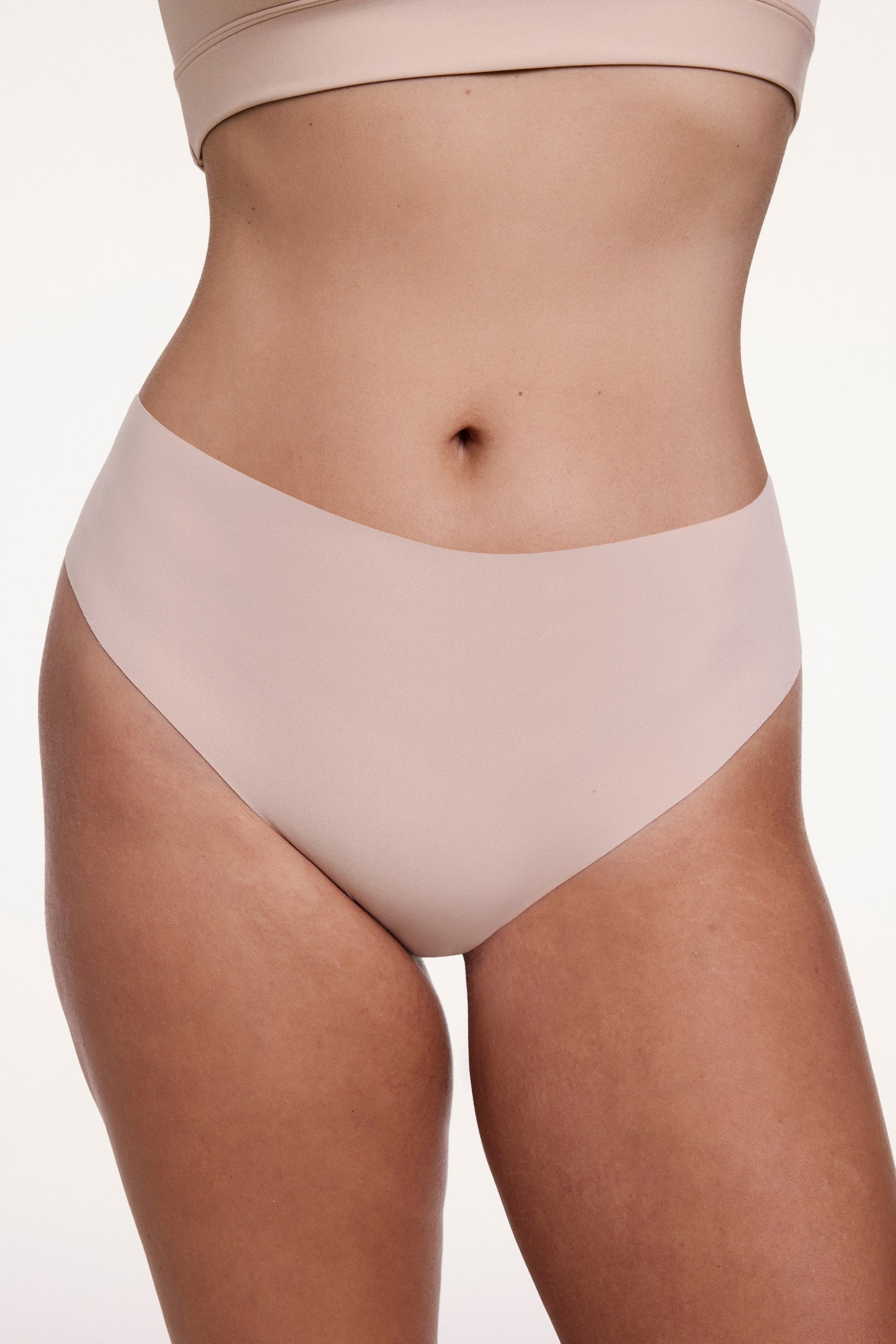 On a white studio backdrop, Sonnie wears the No-Show Highwaist Thong in Beige, shown from the front with a smooth, high-rise waistband and raw-edge finish designed to sit flat against the skin. sizing: Sonnie wears size Large