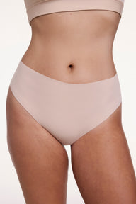 On a white studio backdrop, Sonnie wears the No-Show Highwaist Thong in Beige, shown from the front with a smooth, high-rise waistband and raw-edge finish designed to sit flat against the skin. sizing: Sonnie wears size Large