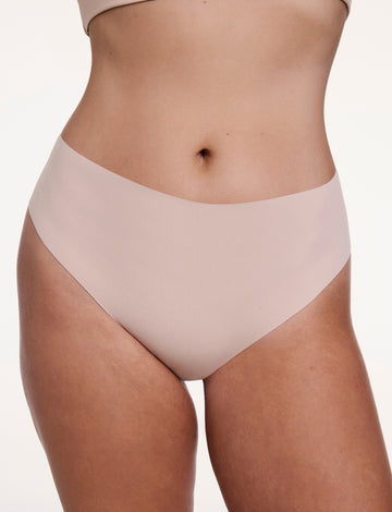 On a white studio backdrop, Sonnie wears the No-Show Highwaist Thong in Beige, shown from the front with a smooth, high-rise waistband and raw-edge finish designed to sit flat against the skin. sizing: Sonnie wears size Large
