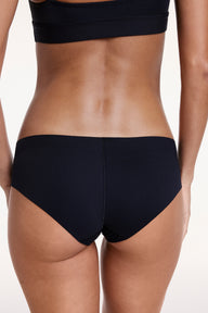 On a white studio backdrop, Linni wears the No-Show Hiphugger in Black, shown from the back with moderate coverage and a single seam designed to sit smoothly under clothing.