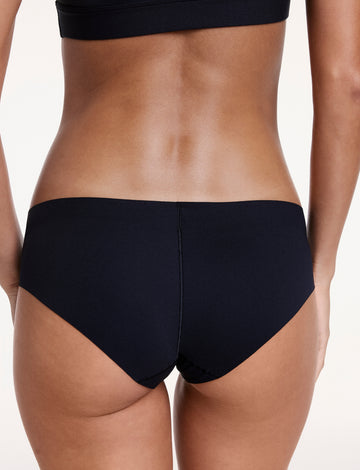 On a white studio backdrop, Linni wears the No-Show Hiphugger in Black, shown from the back with moderate coverage and a single seam designed to sit smoothly under clothing.