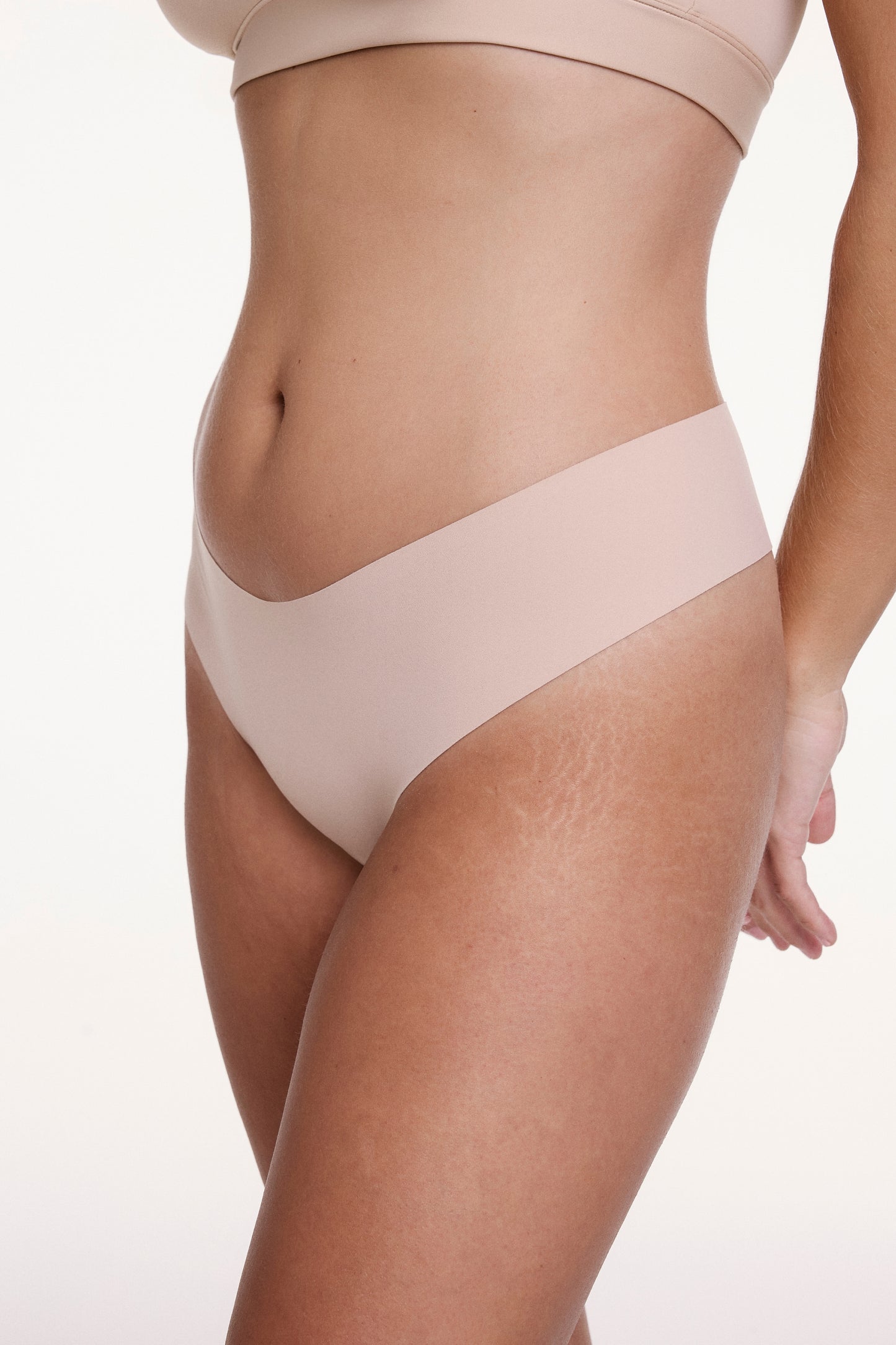 On a white studio backdrop, Sonnie wears the No-Show Low Rise Thong in Beige, shown at an angle to highlight the low-rise fit and smooth, elastic-free edges designed to stay invisible under clothing.
